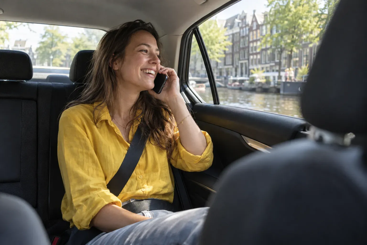 Een vrouw in een geel shirt belt lachend op de achterbank, buiten glijden zonnige grachtengevels voorbij.