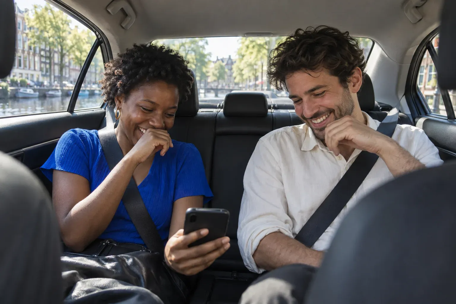 Two friends in the back seat laugh at something on a phone, summer light through the windows.